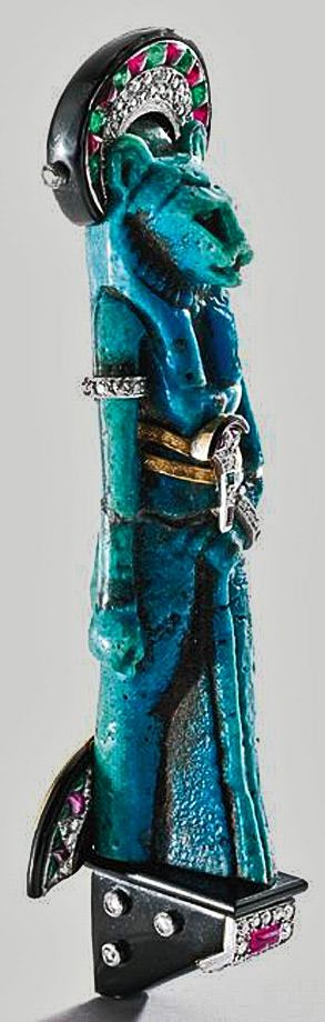 egyptian-revival-sekhmet-brooch Sekhmet-brooch - Egyptian goddess Sekhmet brandishing a fan of stylized lotus blossom motifs, highlighted by a diamond-set bracelet and arm band, fitted at the waist with two gold belts of zig- black enamel. Highlighted by diamonds , rubies and emeralds.